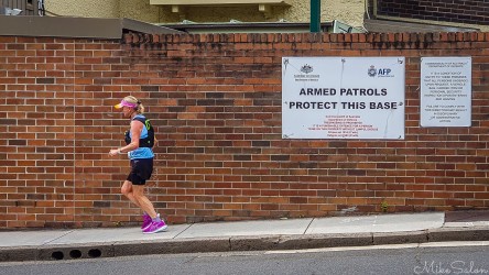 A Bondi‑to‑Manly Ultra runner passes the side wall of Sydney’s Royal Australian Navy base at Woolloomooloo. The warning signs and brick façade mark a sharp contrast to the open harbour views nearby, underscoring the course’s mix of scenic and urban character. [0555]
