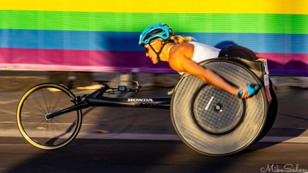 A focused wheelchair athlete gliding past Taylor Square in the early morning sun during the 2025 Sydney Marathon. The rainbow barrier and golden light emphasise speed, strength, and grace in motion. [5198]