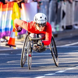 Japanese wheelchair racer Nakam drives hard through Taylor Square during the 2025 Sydney Marathon. The red racing suit and aerodynamic chair cut through the morning light, capturing the precision and power of elite para‑sport competition. [5303]